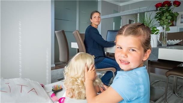 Ein Kind spielt mit einer Puppe, eine Frau sitzt am Schreibtisch