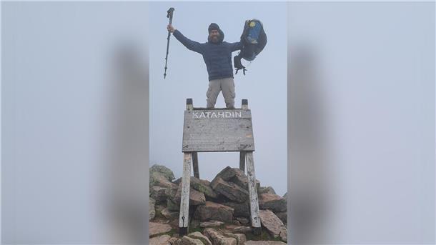 Ein Mann steht am Gipfelkreuz des Mount Katahdin und hält Wanderstöcke und Rucksack in die Luft.