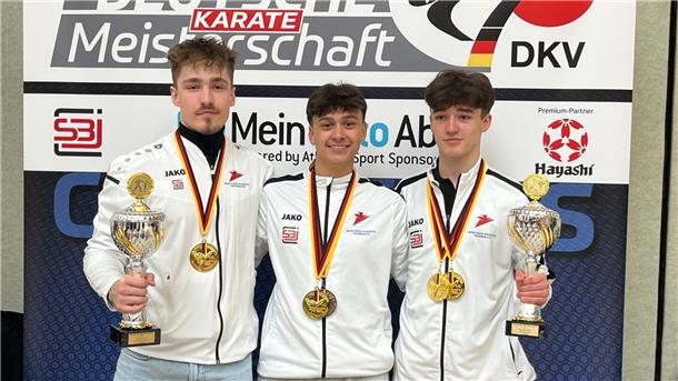Drei Karateka halten Pokale in die Kamera und haben ihre Goldmedaillen um den Hals hängen.