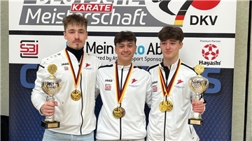 Drei Karateka halten Pokale in die Kamera und haben ihre Goldmedaillen um den Hals hängen.