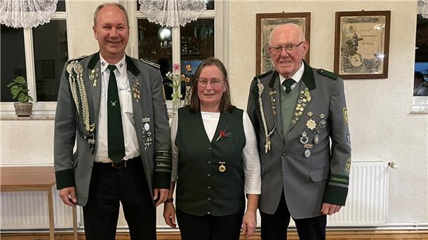 Drei Schützen in traditioneller Uniform stehen nebeneinander und lächeln in die Kamera. Zwei Männer tragen graue Schützenjacken mit Orden, die Frau in der Mitte eine grüne Weste mit Anstecknadel. Im Hintergrund sind Fenster und gerahmte Urkunden zu sehen.