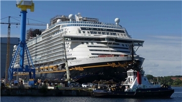 Das Kreuzfahrtschiff „Disney Adventure“ liegt am Werftkai, im Vordergrund ein Schlepper. Am Schiffsrumpf sind Verzierungen und der Schriftzug gut erkennbar.