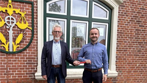Henry Kowalewski und Mathis Jährling stehen vor einem Backsteingebäude und halten gemeinsam symbolisch einen roten Staffelstab – Zeichen des Führungswechsels in der SPD-Fraktion Wurster Nordseeküste.