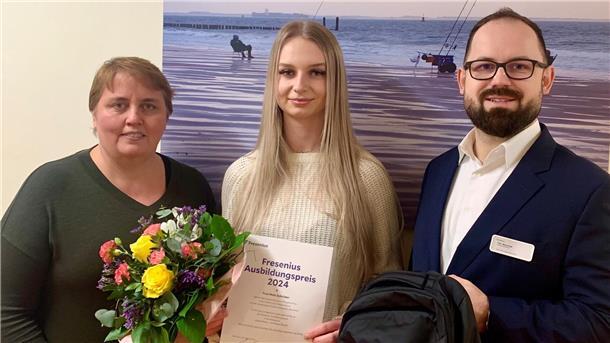 Zwei Frauen und ein Mann stehen vor einer Wand. Sie halten Blumen, eine Urkunde und einen Rucksack in den Händen.