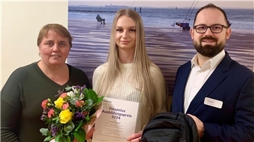 Zwei Frauen und ein Mann stehen vor einer Wand. Sie halten Blumen, eine Urkunde und einen Rucksack in den Händen.