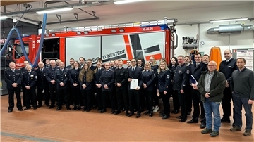 Eine große Gruppe von Feuerwehrleuten in Uniform steht in einer Fahrzeughalle vor einem roten Feuerwehrfahrzeug mit der Aufschrift "Feuerwehr Lunestedt". Einige Personen halten Urkunden oder Dokumente.