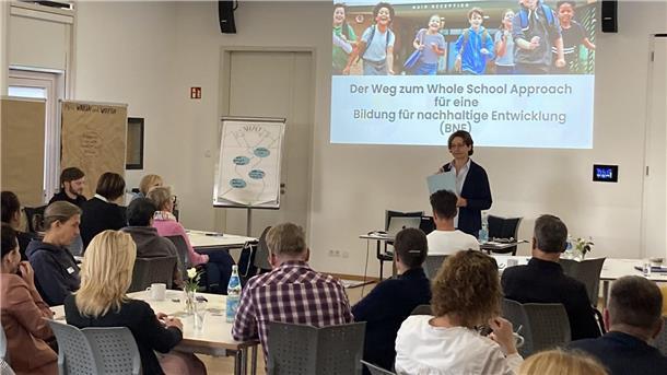Mehrere Menschen sitzen an Tischen und gucken in Richtung einer Frau, die sie über Bildung für eine nachhaltige Entwicklung informiert. 