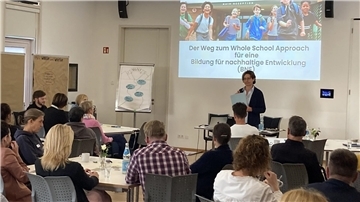 Mehrere Menschen sitzen an Tischen und gucken in Richtung einer Frau, die sie über Bildung für eine nachhaltige Entwicklung informiert. 
