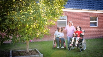 Zwei junge Frauen mit Behinderung stehen mit Ihrem Rollstuhl im Garten vor einer Backsteinwand, ihre Mutter steht zwischen ihnen. Lins im Vordergrund wächst ein Baum. 