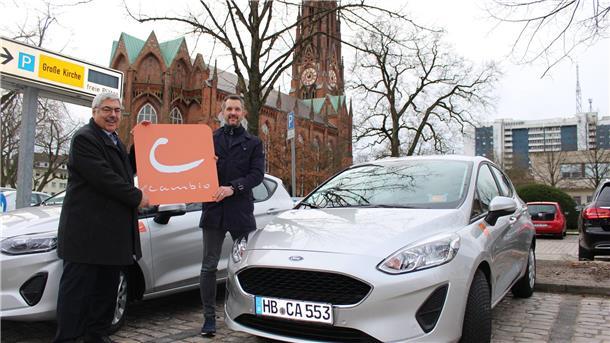 Zwei Männer stehen auf einem Parkplatz vor einem Auto und halten ein Firmenschild in den Händen.