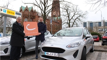Zwei Männer stehen auf einem Parkplatz vor einem Auto und halten ein Firmenschild in den Händen.