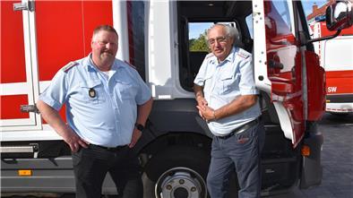 Zwei Feuerwehrmänner in Uniform stehen bei sonnigem Wetter neben einem roten Einsatzfahrzeug der Ortsfeuerwehr Loxstedt. Einer lehnt an der offenen Fahrertür.