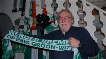 Ein Mann steht vor einem Treppenaufgang mit lauter Fanutensilien. In der Hand hält er einen Schal des SV Werder Bremen.