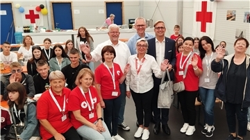 Eine Gruppe von Kindern, Jugendlichen und Erwachsenen winken in die Kamera. Im Hintergrund hängen Flaggen mit dem Symbol des Roten Kreuzes.