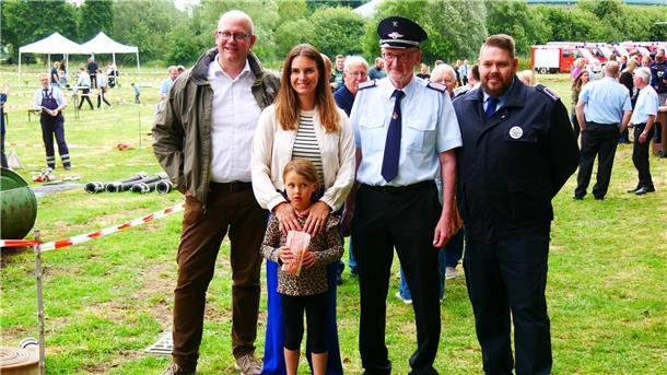 Mehrere Personen, darunter Feuerwehrangehörige in Uniform und eine Politikerin mit Kind und Ehemann, stehen auf einer Wiese beim Feuerwehrfest in Rockstedt. Im Hintergrund laufen Wettkämpfe und Besucher spazieren zwischen Absperrbändern und Einsatzgeräten.