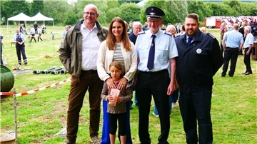 Mehrere Personen, darunter Feuerwehrangehörige in Uniform und eine Politikerin mit Kind und Ehemann, stehen auf einer Wiese beim Feuerwehrfest in Rockstedt. Im Hintergrund laufen Wettkämpfe und Besucher spazieren zwischen Absperrbändern und Einsatzgeräten.