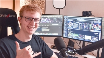 Ein junger Mann mit Brille lächelt in die Kamera. Hinter ihm stehen zwei Computerbildschirme.