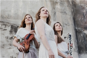Drei Frauen in heller Kleidung (Kleidern) sind zu sehen, die Frau links hat eine Violine in der Hand, die Frau rechts eine Klarinette. Das Trio Luz y Sombra verspricht am 9. Mai einen Abend voller musikalischer Überraschungen.
