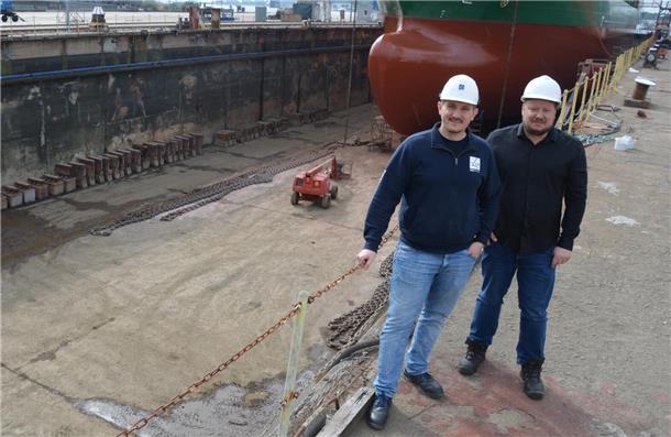 Zwei Männer mit weißen Sicherheitshelmen stehen vor einem großen Trockendock, in dem ein großes Schiff aufgebockt ist. Einer der Männer trägt eine dunkelblaue Jacke, der andere ein schwarzes Hemd. Im Hintergrund ist der Rumpf des Schiffes zu sehen, sowie Industrieketten, Maschinen und die trockene Dockfläche.