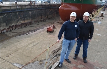 Zwei Männer mit weißen Sicherheitshelmen stehen vor einem großen Trockendock, in dem ein großes Schiff aufgebockt ist. Einer der Männer trägt eine dunkelblaue Jacke, der andere ein schwarzes Hemd. Im Hintergrund ist der Rumpf des Schiffes zu sehen, sowie Industrieketten, Maschinen und die trockene Dockfläche.