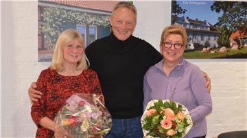 Drei Personen stehen lächelnd vor einer weißen Wand mit Bildern von Gebäuden. Der Mann in der Mitte trägt einen schwarzen Rollkragenpullover und hat seine Arme um die beiden Frauen gelegt. Die Frauen halten Blumensträuße.