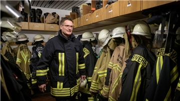 Ein Feuerwehrmann steht in voller Montur in der Umkleidekabine zwischen Feuerwehranzügen. 