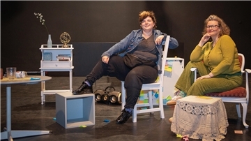 Zwei Personen sitzen auf Stühlen auf einer Theaterbühne.