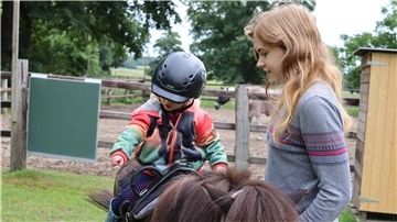 Kind auf Pony, daneben Frau mit langen Haaren