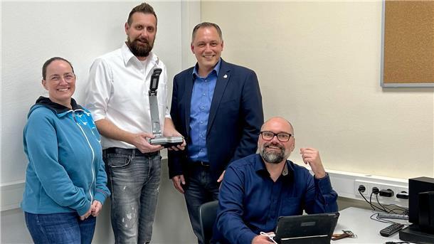 Eine Frau und zwei Männer stehen und sitzen an einem Tisch, ringsherum sind mehrere technische Geräte zu sehen.