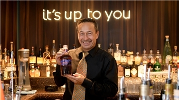 Ein Mann in einer Bar, der seine Rumkreation zeigt. Im Hintergrund ist in leuchtender Schrift "it's up to you" zu lesen. 