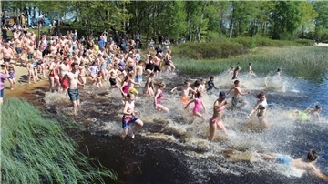 Zahlreiche Menschen, darunter viele Kinder, rennen in Badekleidung bei sonnigem Wetter gemeinsam ins Wasser eines Sees. Am Ufer stehen Zuschauer, im Hintergrund sind Bäume und ein Sandstrand zu sehen.