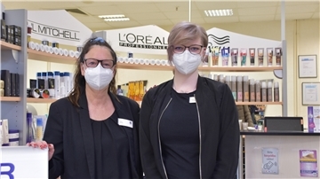 Die beiden Frauen tragen einen Mund-Nasen-Schutz und stehen vor mehreren Friseurutensilien, die in einem Regal liegen.