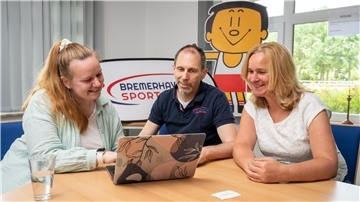 Zwei Frauen und ein Mann sitzen an einem Schreibtisch und blicken auf einen Laptop. Im Hintergrund ist das Logo der Bremerhavener Sportjugend zu sehen.