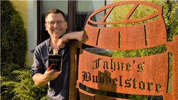 Ein Mann steht neben einem Schild. Es weist auf den Rumshop Fahne's Buddelstore von Sascha Fähndrich hin. In seiner rechten Hand hält der Mann eine Wagemut-Rumflasche. 