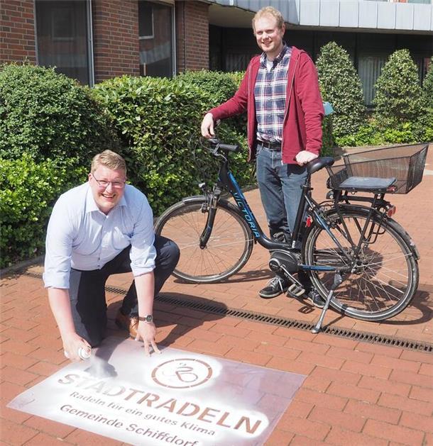 Schiffdorfs Bürgermeister Henrik Wärner sprüht per Schablone Kreide auf den Zugang zum Rathaus. Klimaschutzmanager Birger Auffahrt steht mit einem Fahrrad hinter ihm.