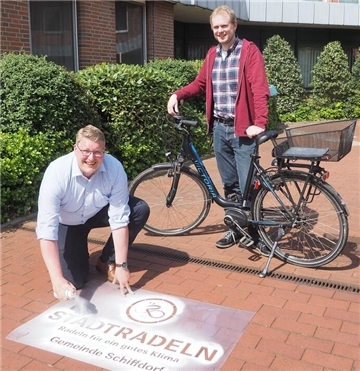 Schiffdorfs Bürgermeister Henrik Wärner sprüht per Schablone Kreide auf den Zugang zum Rathaus. Klimaschutzmanager Birger Auffahrt steht mit einem Fahrrad hinter ihm.