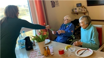 eine Frau überreicht zwei Seniorinnen eine Rose über den Frühstückstisch