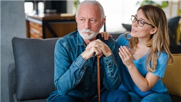 Ein älterer Herr und eine junge Frau sitzen auf einem Sofa, wobei er offensichtlich nicht hört, was sie sagt.
