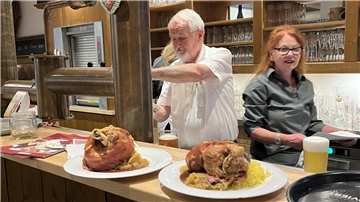 Ein älterer Herr in weißem Hemd steht am Tresen und zapft Bier. Vor ihm stehen zwei Teller mit Riesenhaxe. Eine Dame geht am Mann vorbei und hält Zettel in der Hand. 