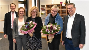 Fünf Personen nebeneinander. Links ein Mann, rechts daneben drei Frauen, zwei halten einen Blumenstrauß in den Händen und ganz rechts steht ein Mann.