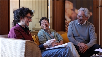 Drei Personen sitzen gemeinsam auf einem Sofa, lachen und unterhalten sich angeregt. Sie wirken entspannt und fröhlich, vor ihnen liegen Notizen und Papiere.