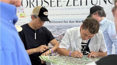 Zwei Männer stehen vor einem Poster, das auf einem hohen Tisch liegt. Einer von ihnen zeichnet darauf.