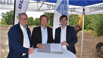 Drei Männer stehen an einem Tisch und zeigen eine Vereinbarung.