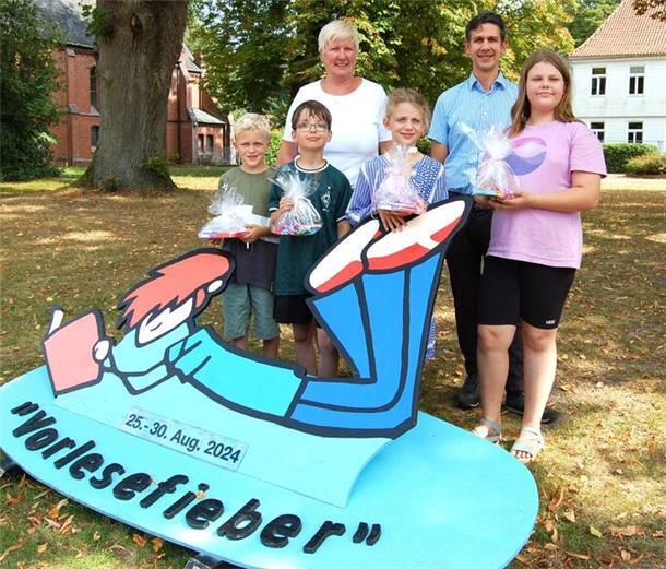 Zwei Erwachsene und vier Kinder mit Geschenken stehen auf einer Grünfläche vor einem Aufsteller, auf dem "Vorlesefieber" und "25.-30. Aug. 2024" steht.