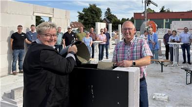 Eine Frau kippt einen Eimer mit Beton in eine Vorrichtung. Gegenüber steht ein lächelnder Mann in Hemd mit einer Kelle voller Beton.