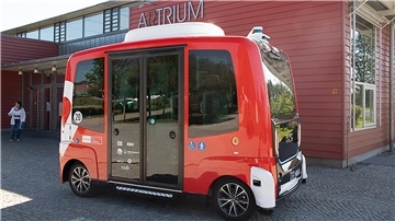 Ein rot-weißer autonomer Kleinbus steht vor einem Gebäude mit der Aufschrift „ARTRIUM“ an der Fassade. Der futuristisch wirkende Bus ohne Fahrerplatz ist mit Symbolen für Barrierefreiheit und Fußgänger gekennzeichnet.