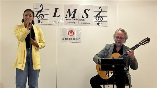 Eine junges Mädchen singt in ein Mikrofon, während ein älterer Mann sie auf einer klassischen Gitarre begleitet. Im Hintergrund hängen Schilder mit der Aufschrift „LMS – Loxstedter Musikschule“ sowie das Logo von „Jugend musiziert“.