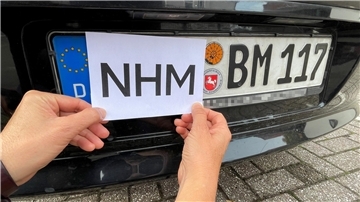 Eine Frau hält einen Zettel mit der Aufschrift NHM auf ein Autokennzeichen.
