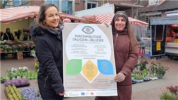 Sophie Schad und Katja Beier präsentieren ein Plakat für die Veranstaltungsreihe "Nachhaltige Augenblicke"