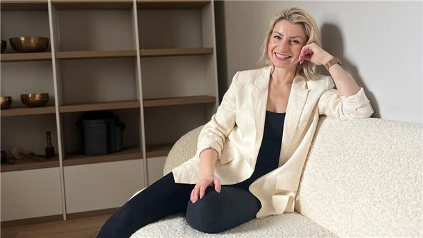 Eine Frau mit blonden Haaren, die einen weißen Blazer und eine dunkle Hose trägt, sitzt auf einer weißen Couch.